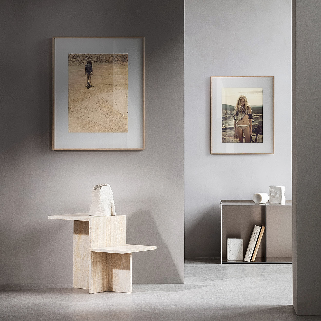 Two framed photographs on a gray wall, with a pale sculptural chair and minimal objects on a low shelf below.