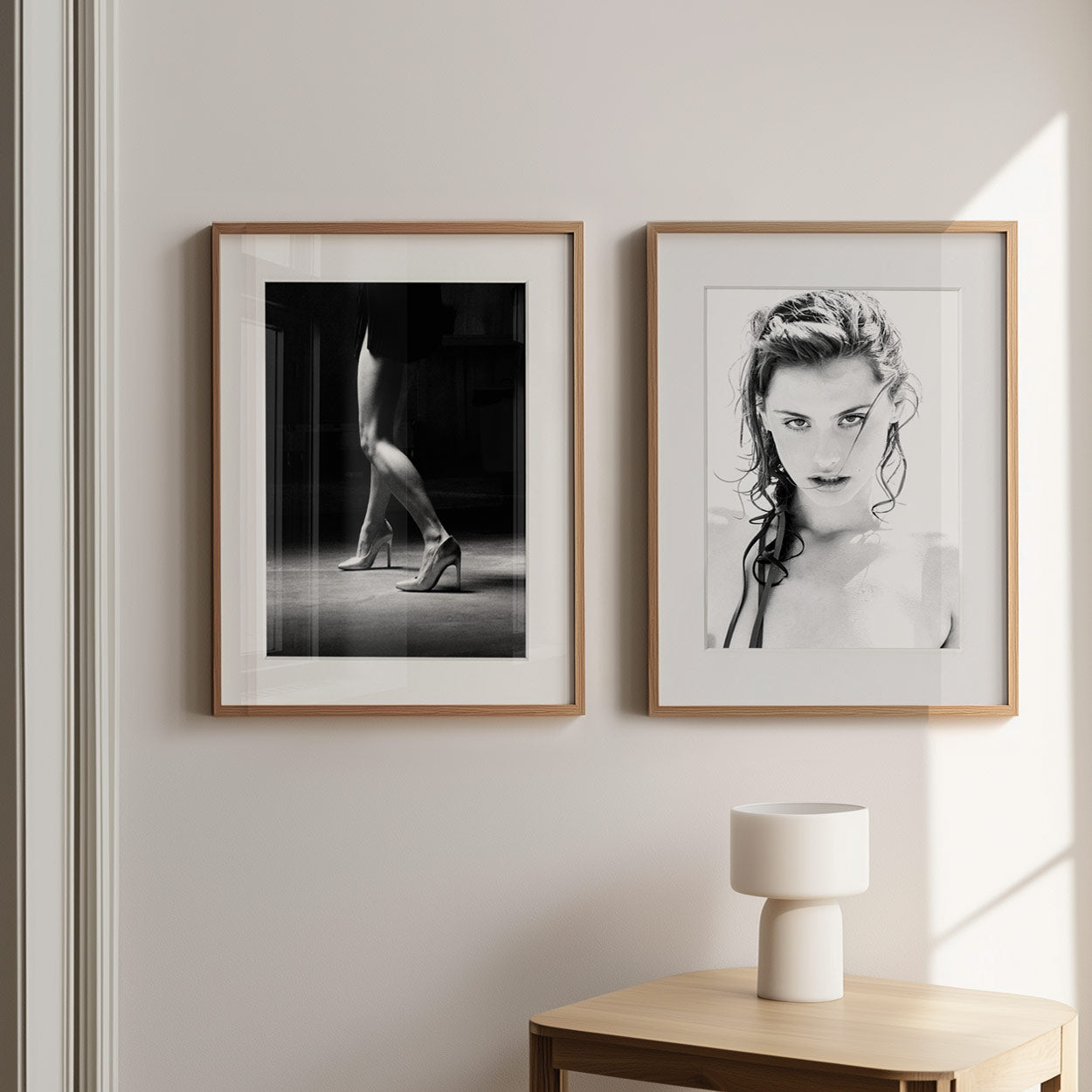 Two framed black-and-white photographs hang on a pale wall above a small wooden table with a white lamp in sunlight.
