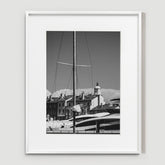 A sailboat mast rises in front of docked yachts and a row of buildings with a clock tower under a clear sky.