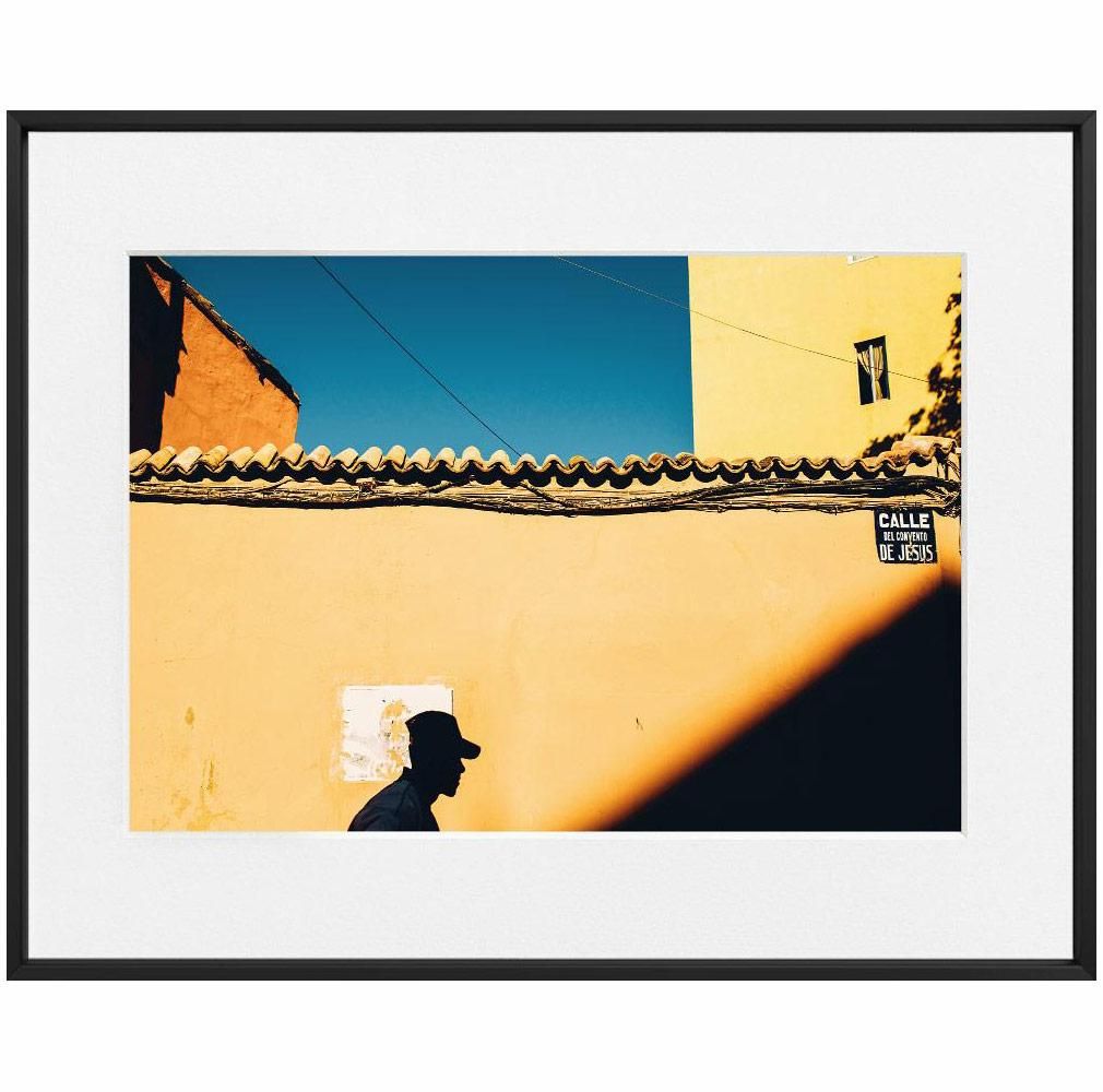 A dark silhouette of a person in profile crosses in front of a sunlit yellow wall beneath a deep blue sky and tiled roofline.