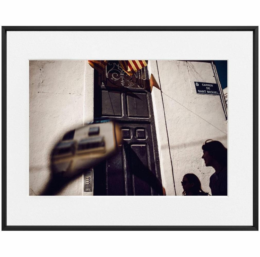 A dark doorway and a street sign reading 'Carrer de Sant Miquel' on a white wall, with two silhouetted figures in the foreground.