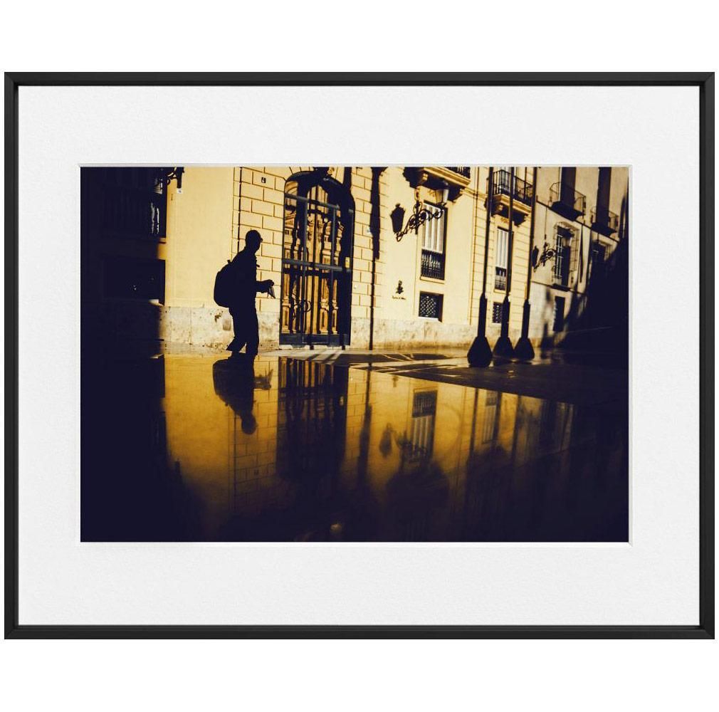 A silhouetted figure with a backpack is reflected on a wet pavement in front of a sunlit building with arched doors and windows.