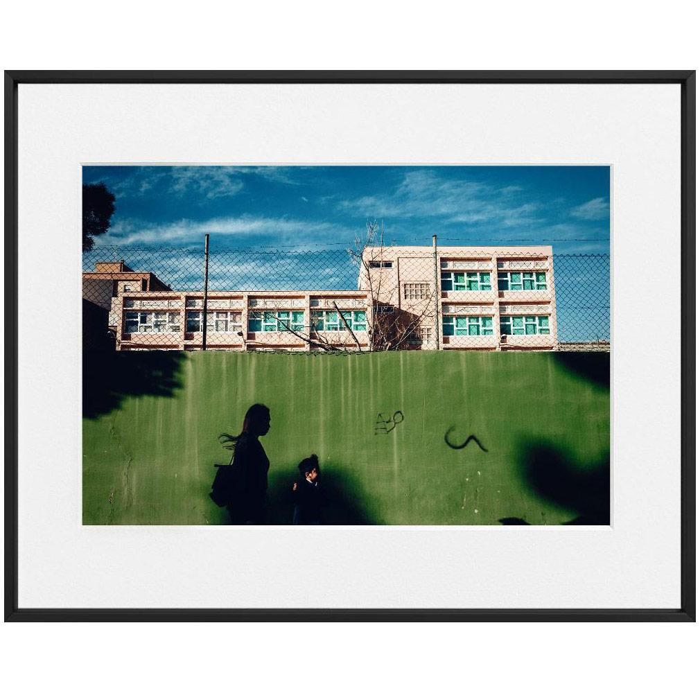 A green wall with graffiti runs across the foreground, casting shadows of two figures below a building with turquoise window frames.