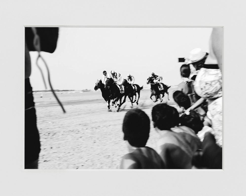 Three jockeys race horses along a sandy stretch as a crowd watches from the right foreground, some holding up cameras or phones.