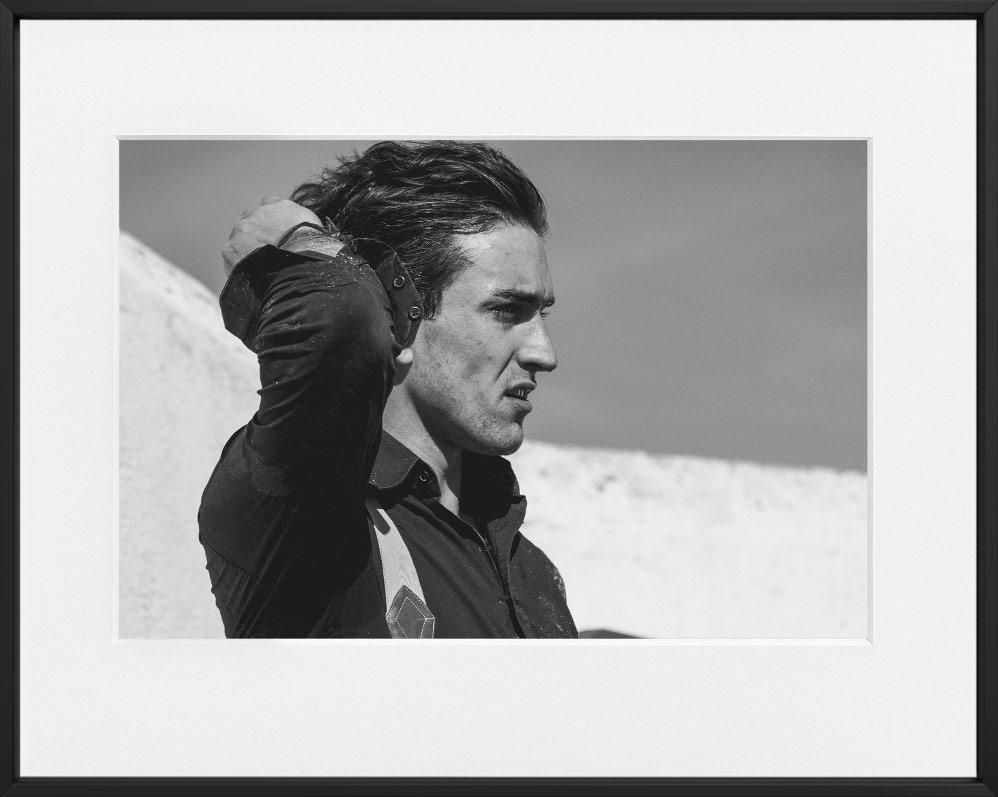 A man in a dark shirt raises one arm to his head, standing outdoors against a pale wall and clear sky.