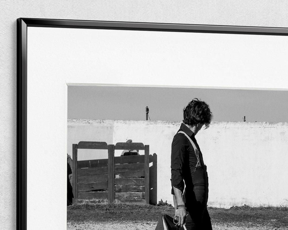 A person with curly hair and rolled sleeves holds an object while walking past a white wall and wooden gate under a clear sky.