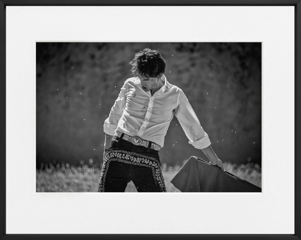 A person in a decorated belt and patterned trousers holds a cloth, standing outdoors with small particles visible in the sunlight.