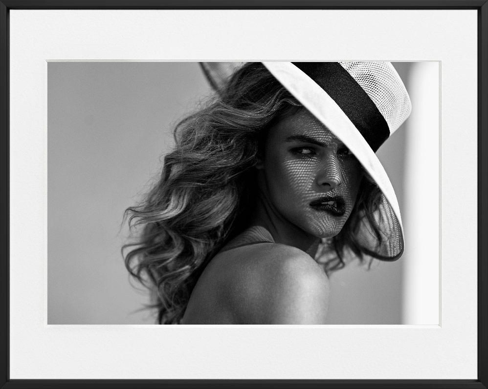 A person with wavy hair wears a wide-brimmed hat with a dark band, casting a mesh shadow pattern across their face.