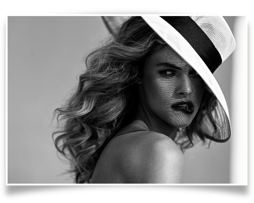 A person with wavy hair wears a wide-brimmed hat, with mesh shadows cast across their face against a plain background.