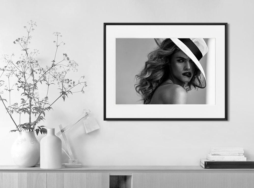 A framed portrait of a woman in a wide-brimmed hat hangs above a light wood cabinet with vases, a lamp, and stacked books.