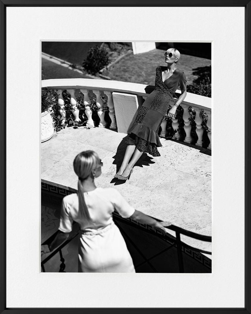 Two women on a sunlit terrace, one leaning against a balustrade in patterned dress, the other in white viewed from behind.