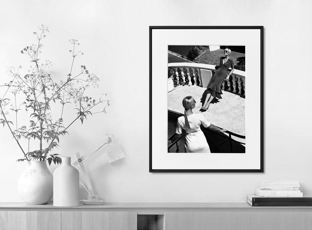 Framed black-and-white photograph on a wall above a light wood cabinet, with a vase of tall flowers and a desk lamp nearby.