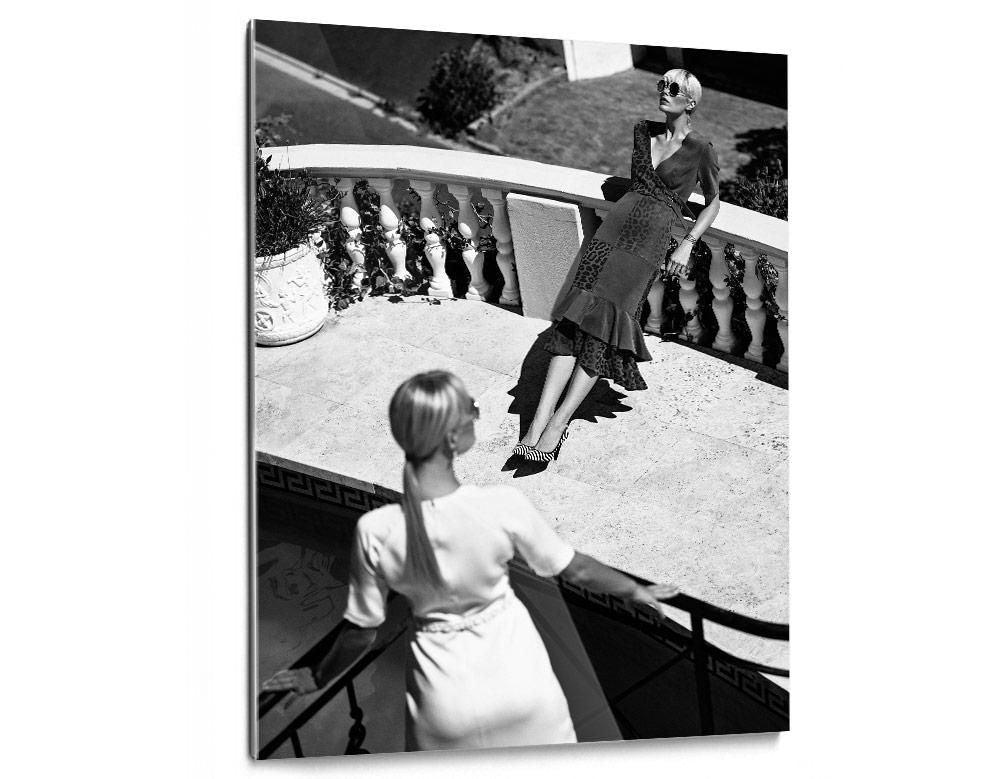 Two women on a sunlit terrace, one leaning against a balustrade in patterned dress, the other seen from behind descending stairs.