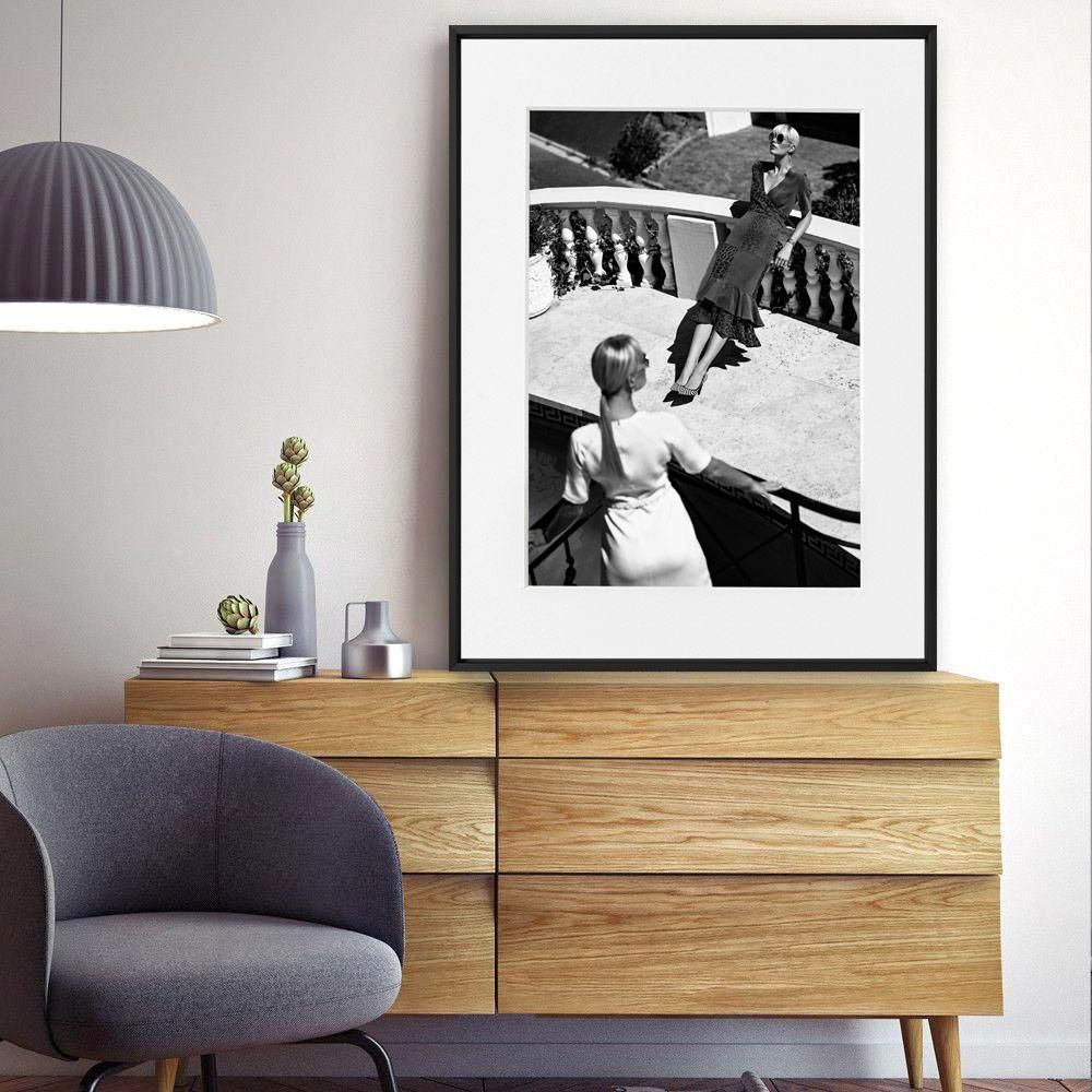 A framed black-and-white photograph of two women on a terrace hangs above a wooden sideboard with vases and stacked books.