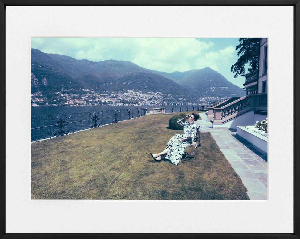 A woman in a patterned dress sits on a bench beside a lawn, with mountains and water visible in the background under a partly cloudy sky.