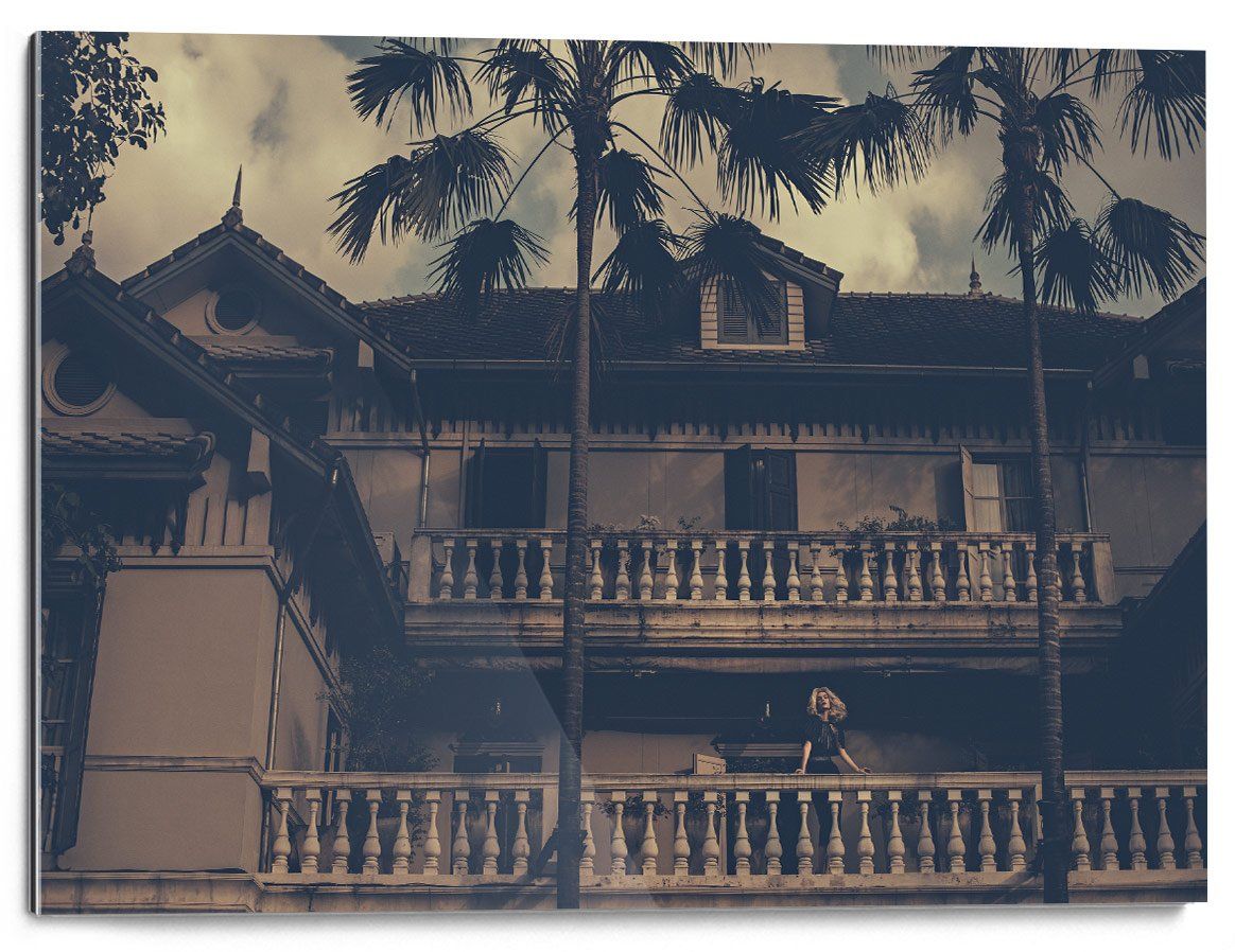 Tall palm trees partially obscure a two-story house with a tiled roof and balustrades; a person is visible on the lower balcony.