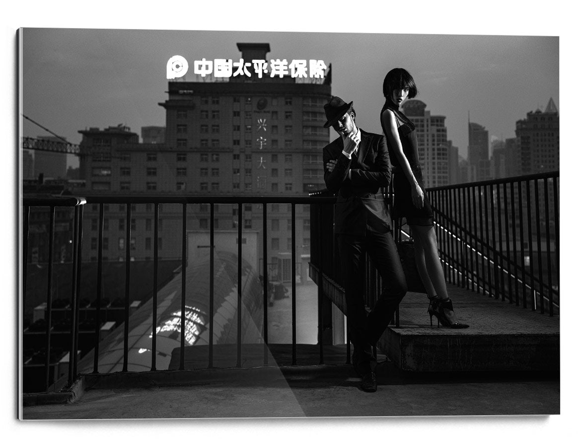 Two people in dark clothing stand on a rooftop stair landing, with a city skyline and a large building in the background.