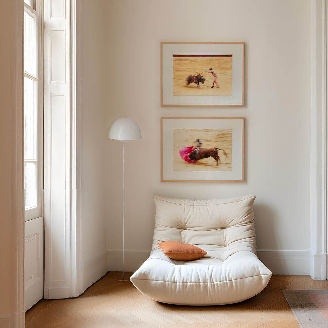 A cream-colored lounge chair with an orange cushion sits beside a tall white lamp under two framed blurred bullfighting prints.
