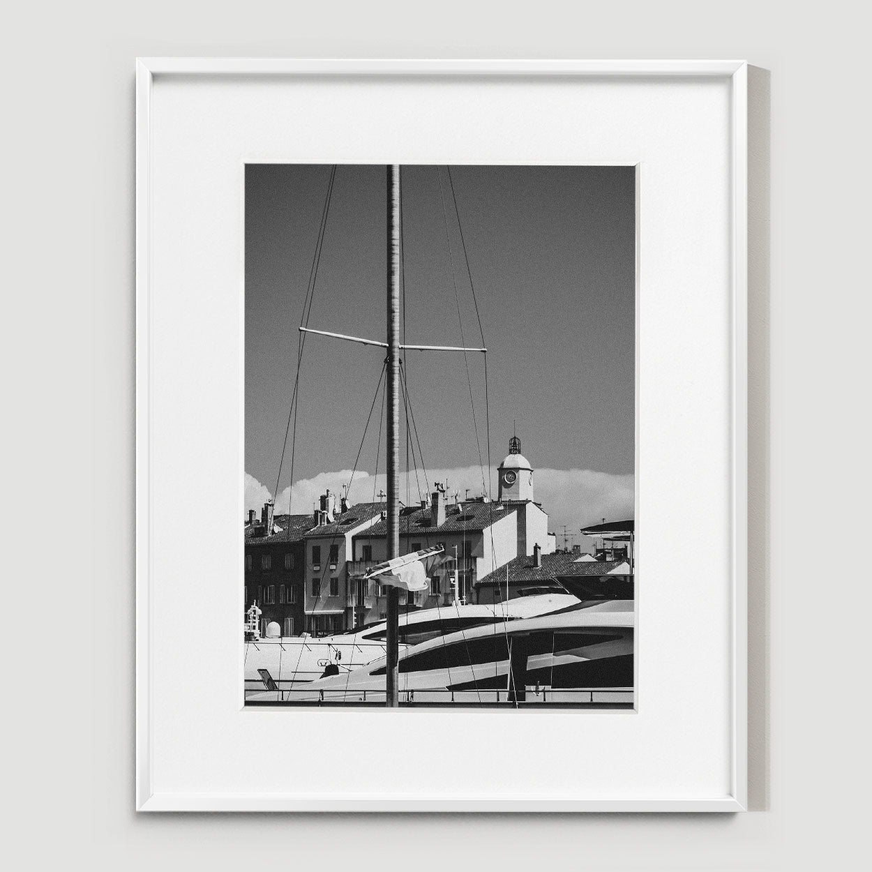 A sailboat mast rises in front of docked yachts and a row of buildings with a clock tower under a clear sky.
