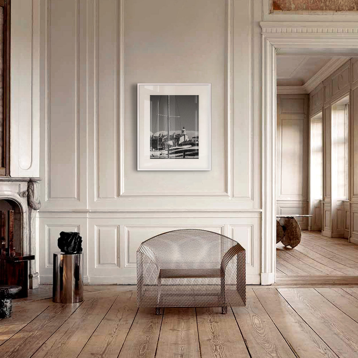 A metal mesh chair sits on wide wooden floorboards in a pale paneled room, with a framed black-and-white print on the wall above.
