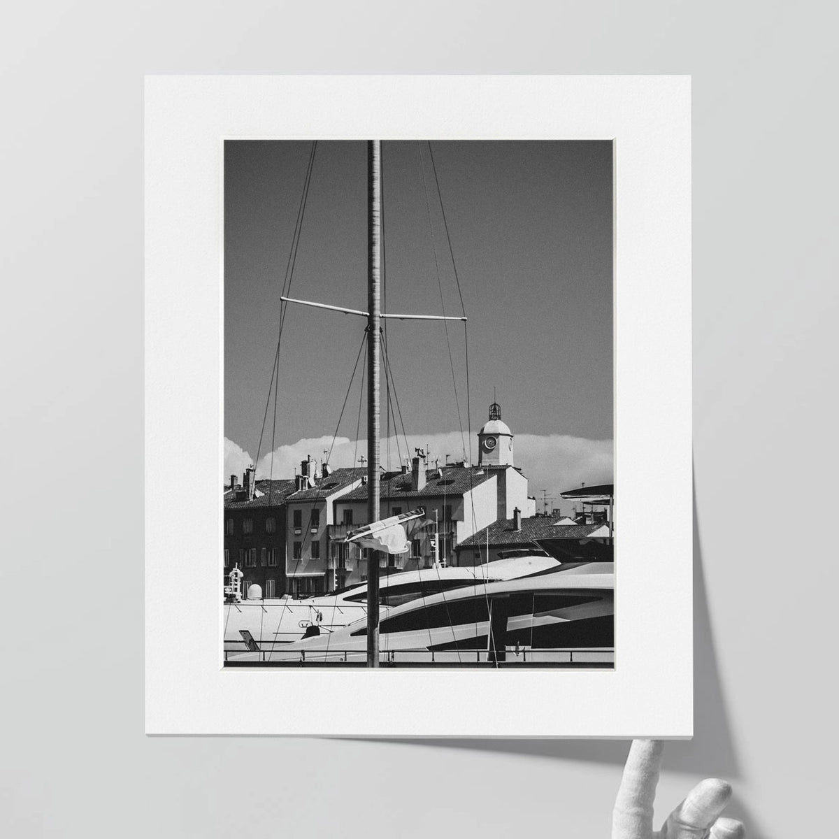A sailboat mast rises in front of docked boats and buildings with a clock tower under a clear sky.