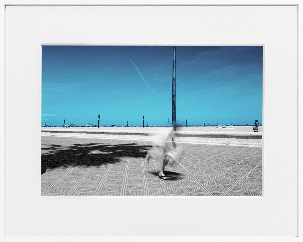A limited edition gallery-quality photograph featuring a person in motion, possibly skateboarding, on a street with a clear blue sky in the background. 