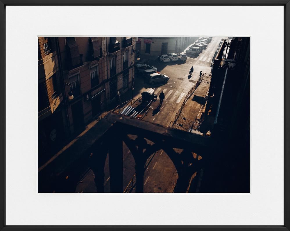 Sunlight casts long shadows across a city street with parked cars, a few figures walking, and buildings lining both sides.