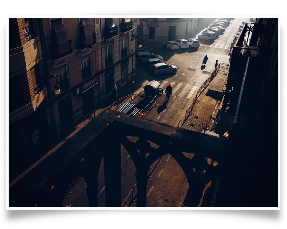 Sunlit street viewed from above, with parked cars, a few figures crossing, and long shadows cast along the pavement and buildings.
