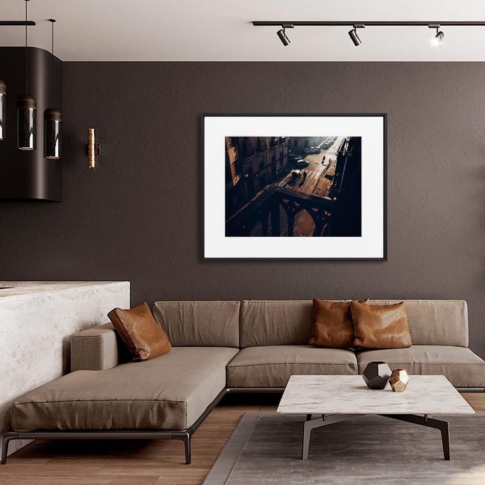 A framed aerial street scene with long shadows hangs above a taupe sectional sofa and a marble coffee table in a modern living room.