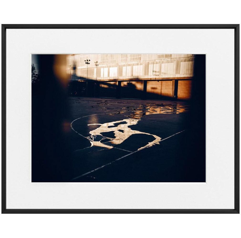 Puddles reflecting a sunlit building stretch across the surface of an empty outdoor court marked with white lines.