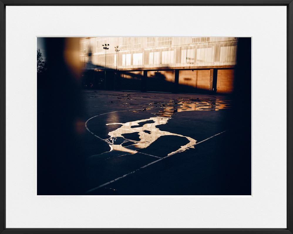 Sunlight reflects off irregular puddles on a dark outdoor court, with a building’s facade mirrored in the water.