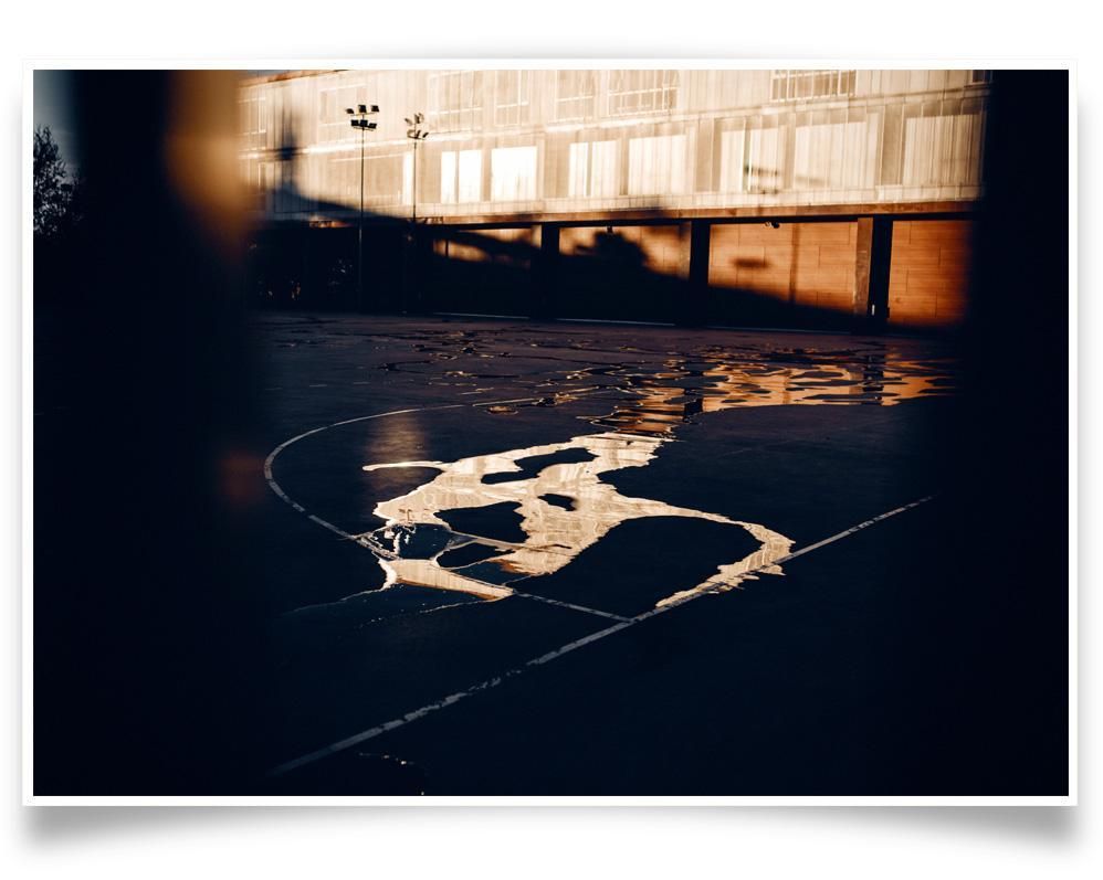 Puddles reflecting warm light stretch across the surface of an outdoor court, with a building and shadows in the background.