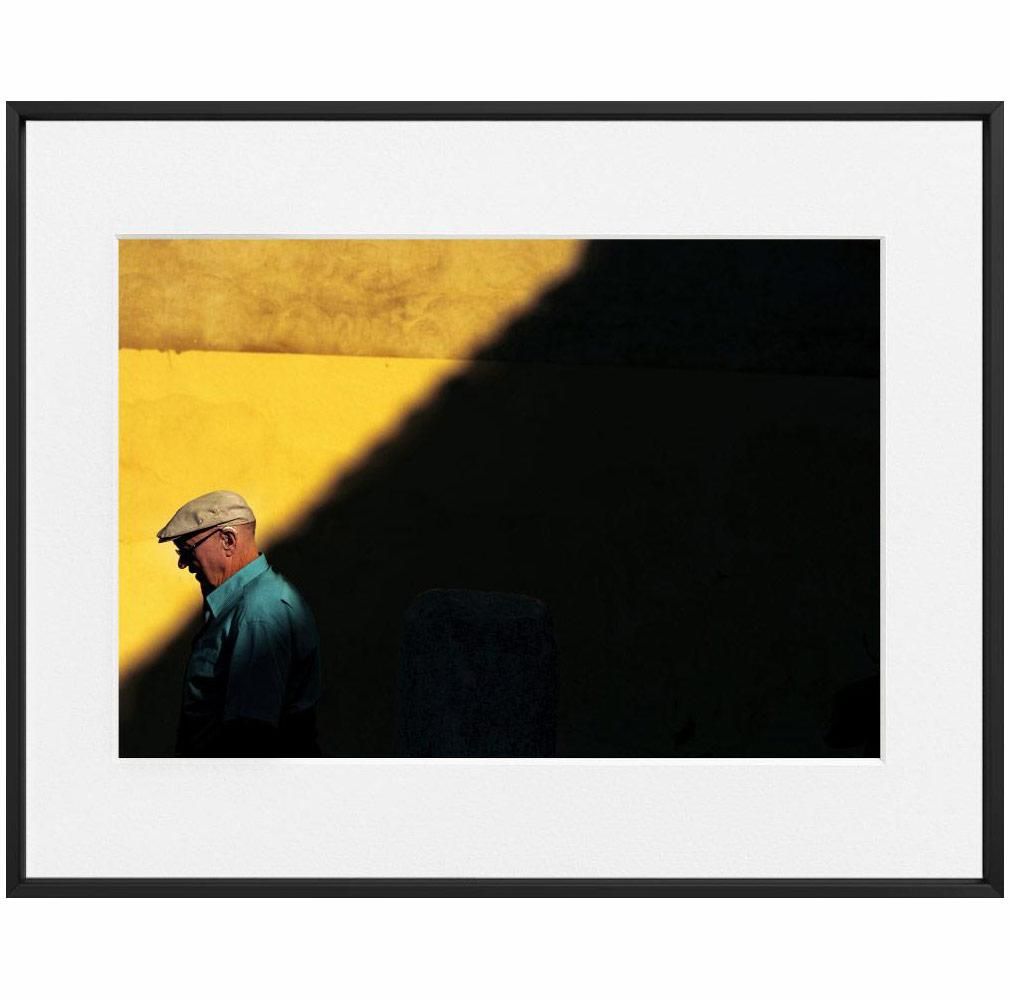 A man in a cap passes through a band of sunlight against a yellow wall, with deep shadow cutting diagonally across the scene.
