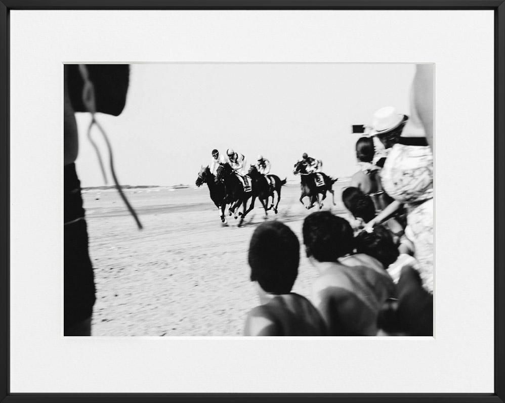Spectators watch three jockeys racing horses along a sandy shoreline, crowd gathered closely in foreground.