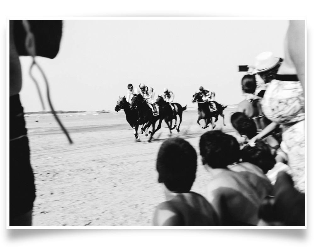 Three jockeys race horses along a sandy shoreline as a crowd of spectators watches from the foreground. The scene is captured in black and white.
