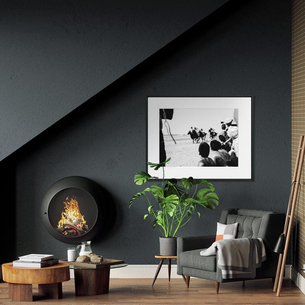 A dark-walled living room with a round wall-mounted fireplace, green potted plant, low wooden tables, and a framed black-and-white photo above.
