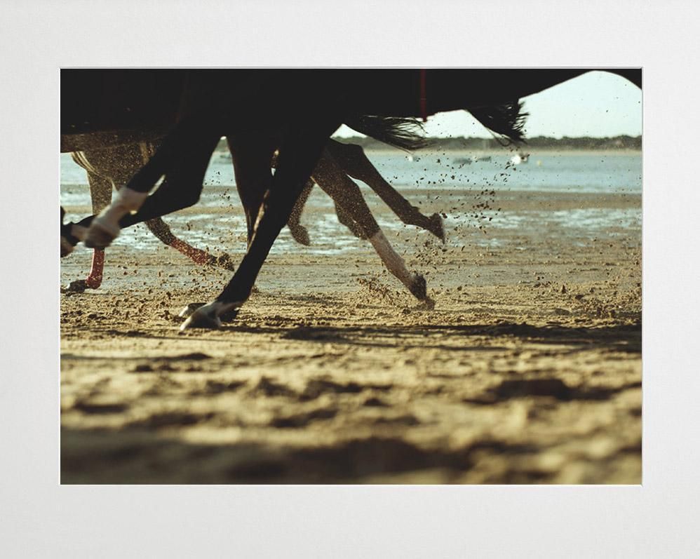 Horse legs in motion kick up sand along a beach, with water and a distant shoreline in the background.
