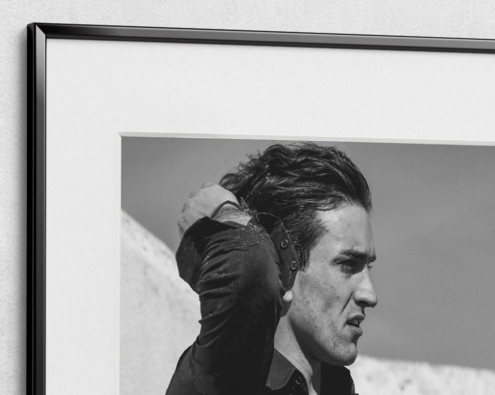 A man with dark hair combed back raises his arm, shown in profile against a plain outdoor background.