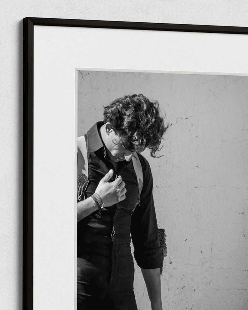A person with curly hair, wearing a dark shirt and suspenders, stands against a textured, light-colored wall.