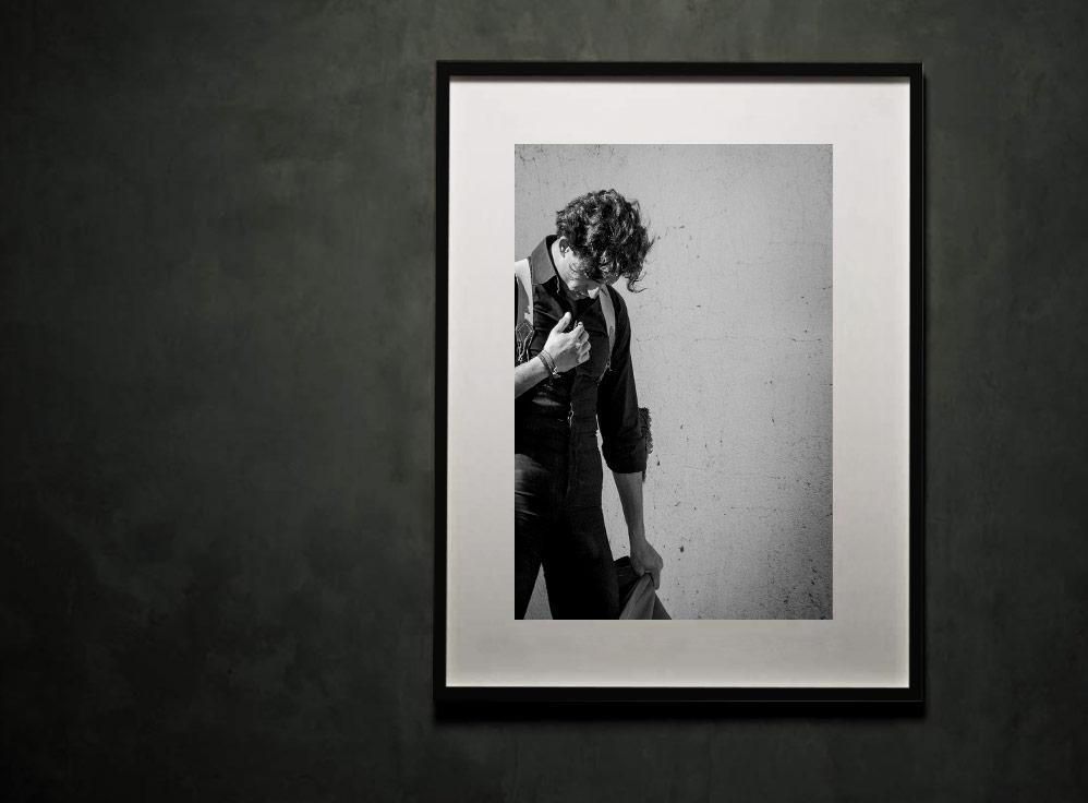 A framed black-and-white portrait shows a person with curly hair looking downward against a plain, textured wall.