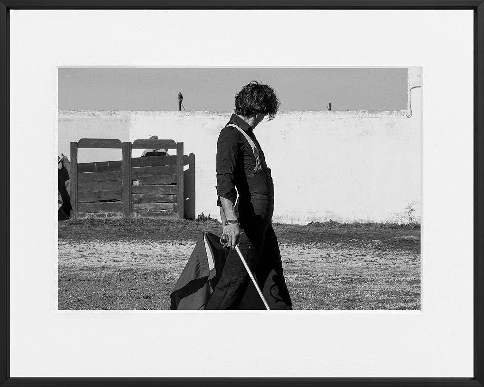 A person in dark clothing walks across a dirt lot, holding a pole and cloth, with a white wall and wooden gate in the background.