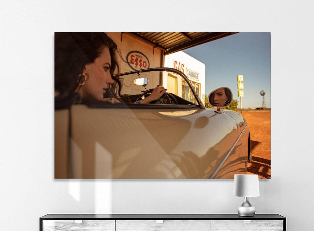 A woman sits in a vintage convertible at a gas station, her reflection visible in the side mirror under a clear sky.