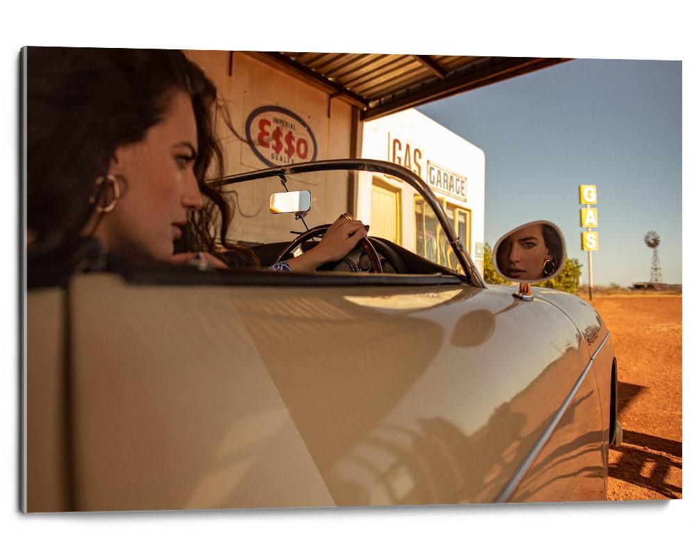 Two women in a tan convertible parked beside a gas station, with one face reflected in the side mirror and a clear blue sky overhead.