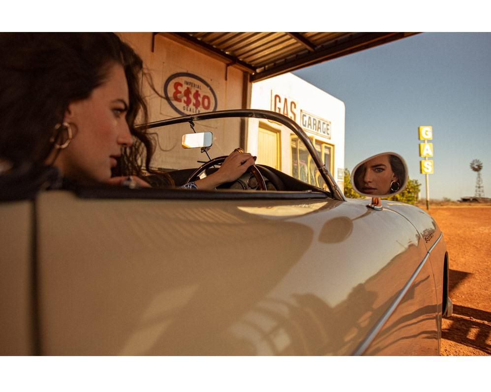 Two women sit in a convertible parked beside a gas station under a metal awning, with a bright sky and a windmill in the distance.