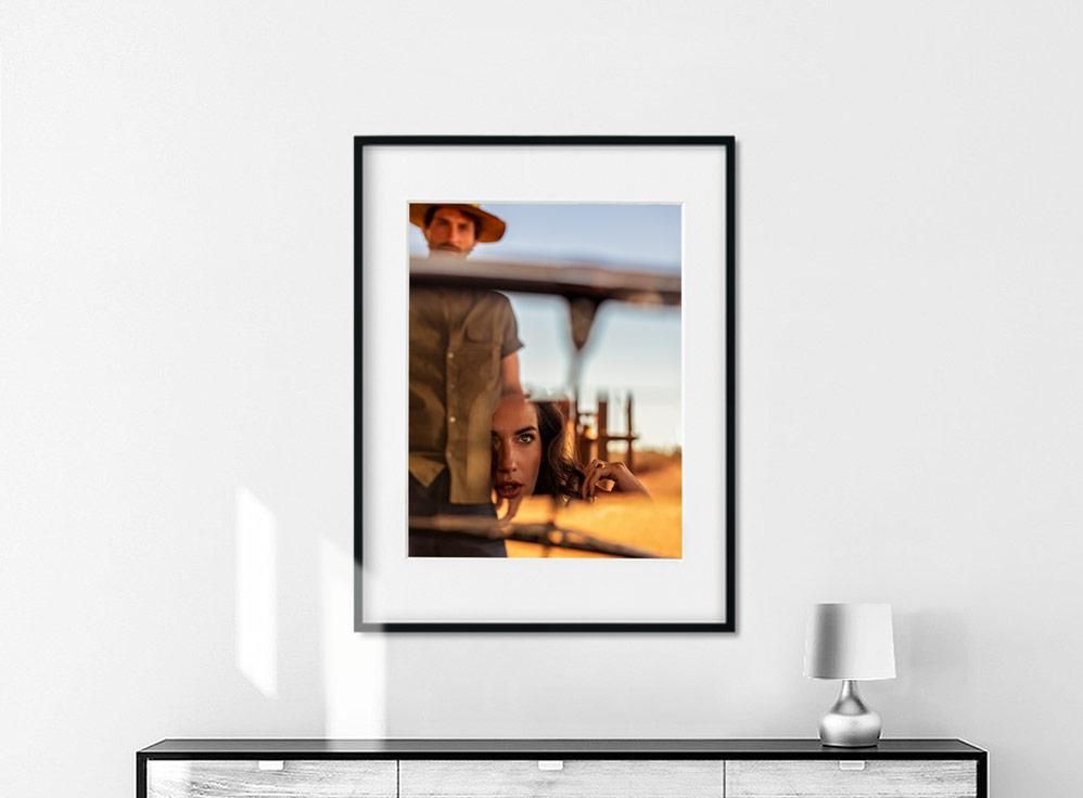 Framed photograph on a white wall above a cabinet, showing two people outdoors with one reflected in glass.