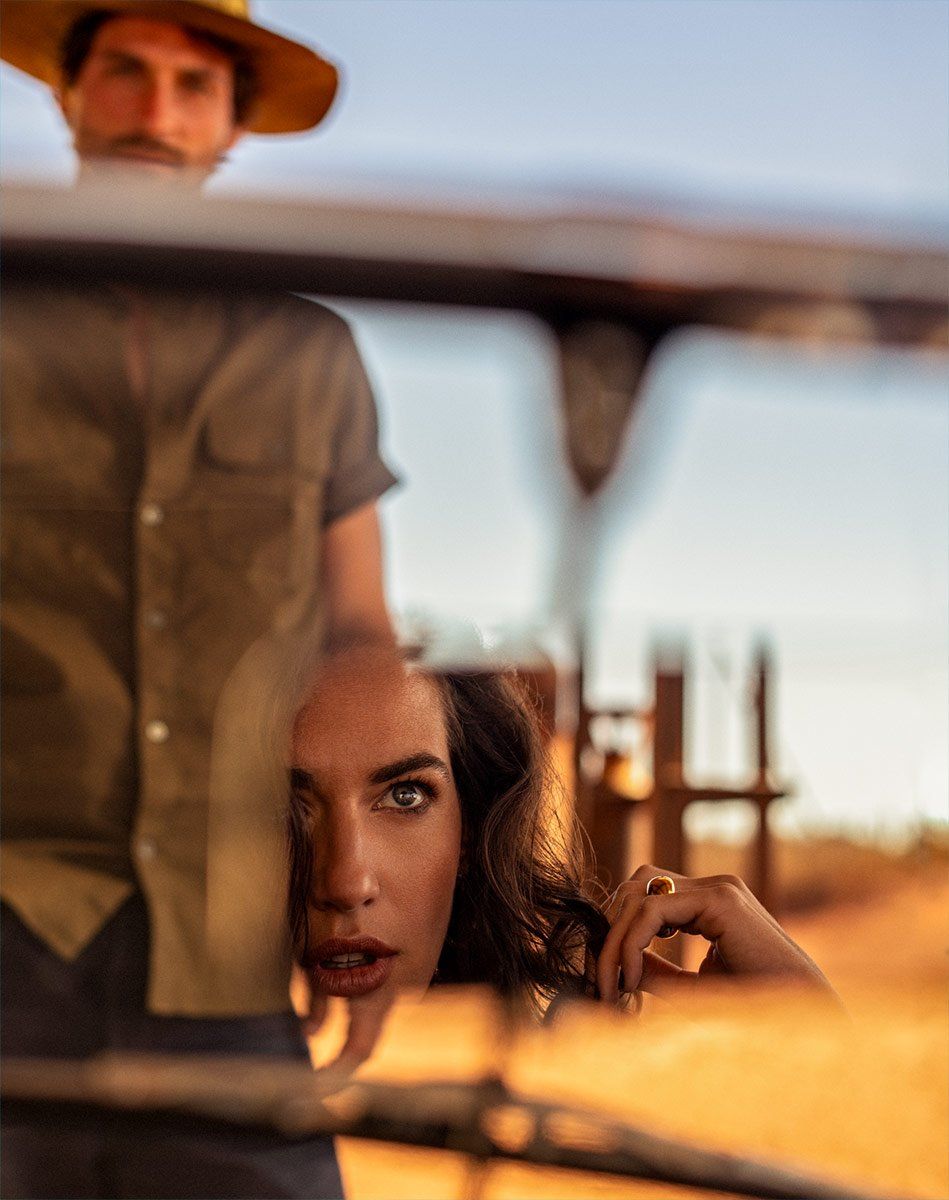 A woman's face and hand are reflected in glass, with a man in a hat partially visible in the blurred background outdoors.