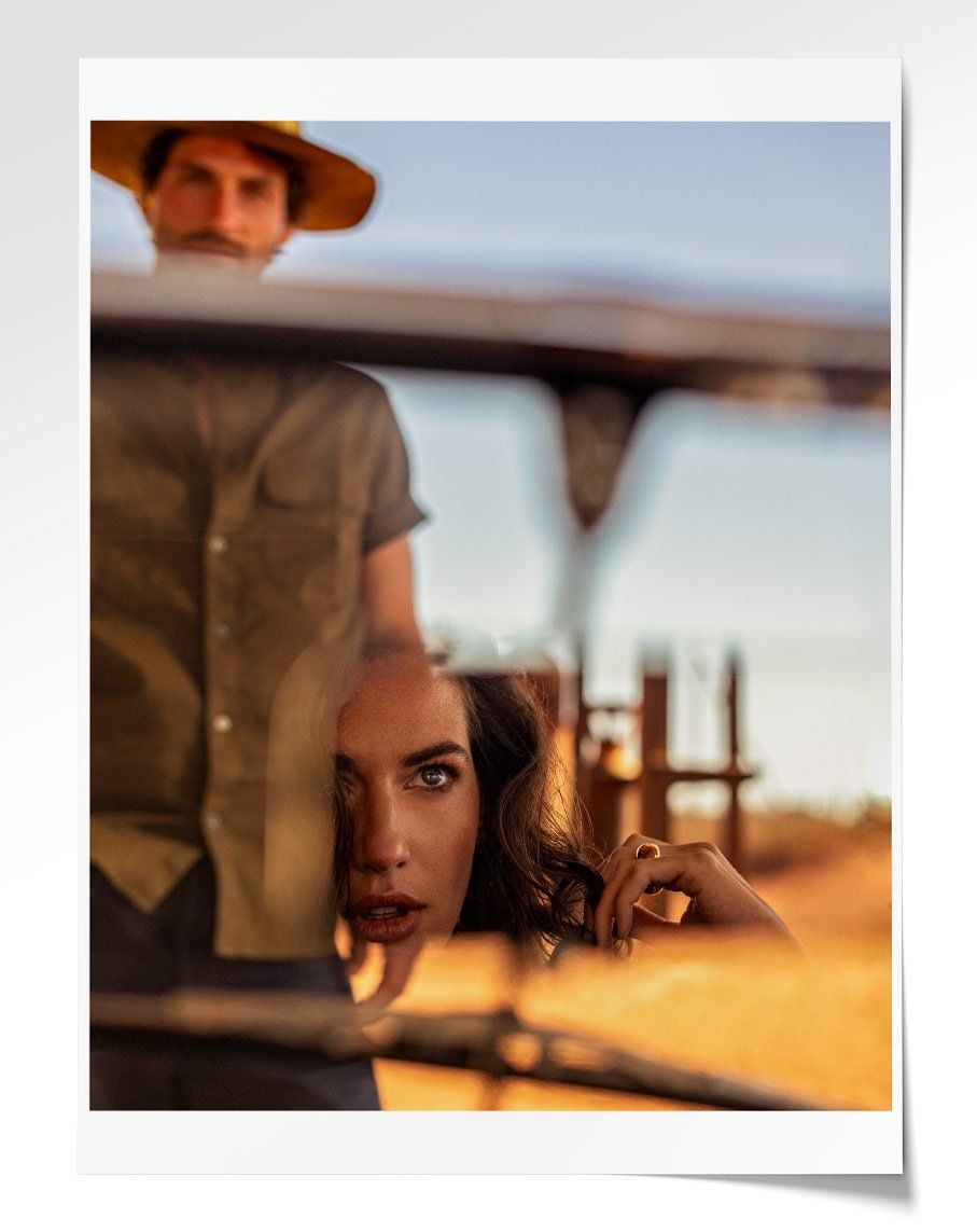 A man in a wide-brimmed hat appears blurred in the background, while a woman's face and hand are reflected sharply in the foreground glass.
