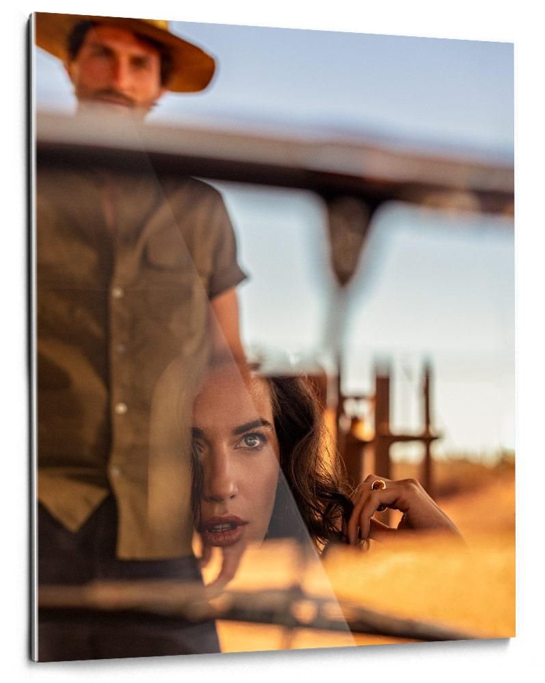 A man in a hat stands behind a woman whose face is reflected in a window, with warm sunlight and blurred outdoor structures in the background.
