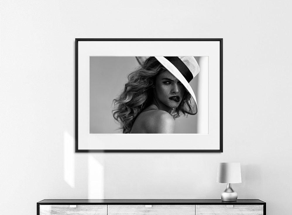 Framed portrait of a person in a wide-brimmed hat with loose hair, mounted above a low cabinet and next to a small table lamp.