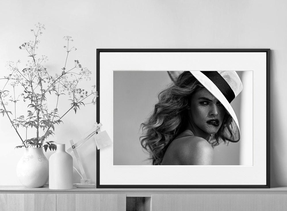 A framed portrait of a person in a wide-brimmed hat rests on a light wooden surface beside vases and a sprig of delicate flowers.